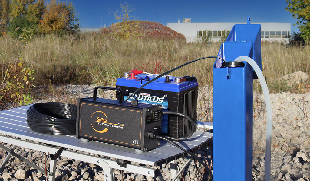 bomba sumergible solinst de 12v en una mesa de campo, conectada a una batería marina de 12v junto a un pozo de monitoreo azul